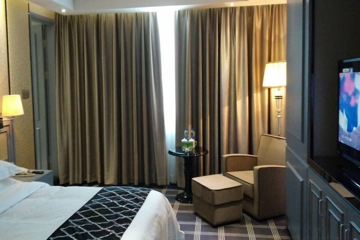 Habitación de hotel con cama, televisión, silla, una mesita con lámpara y ventana con cortinas.