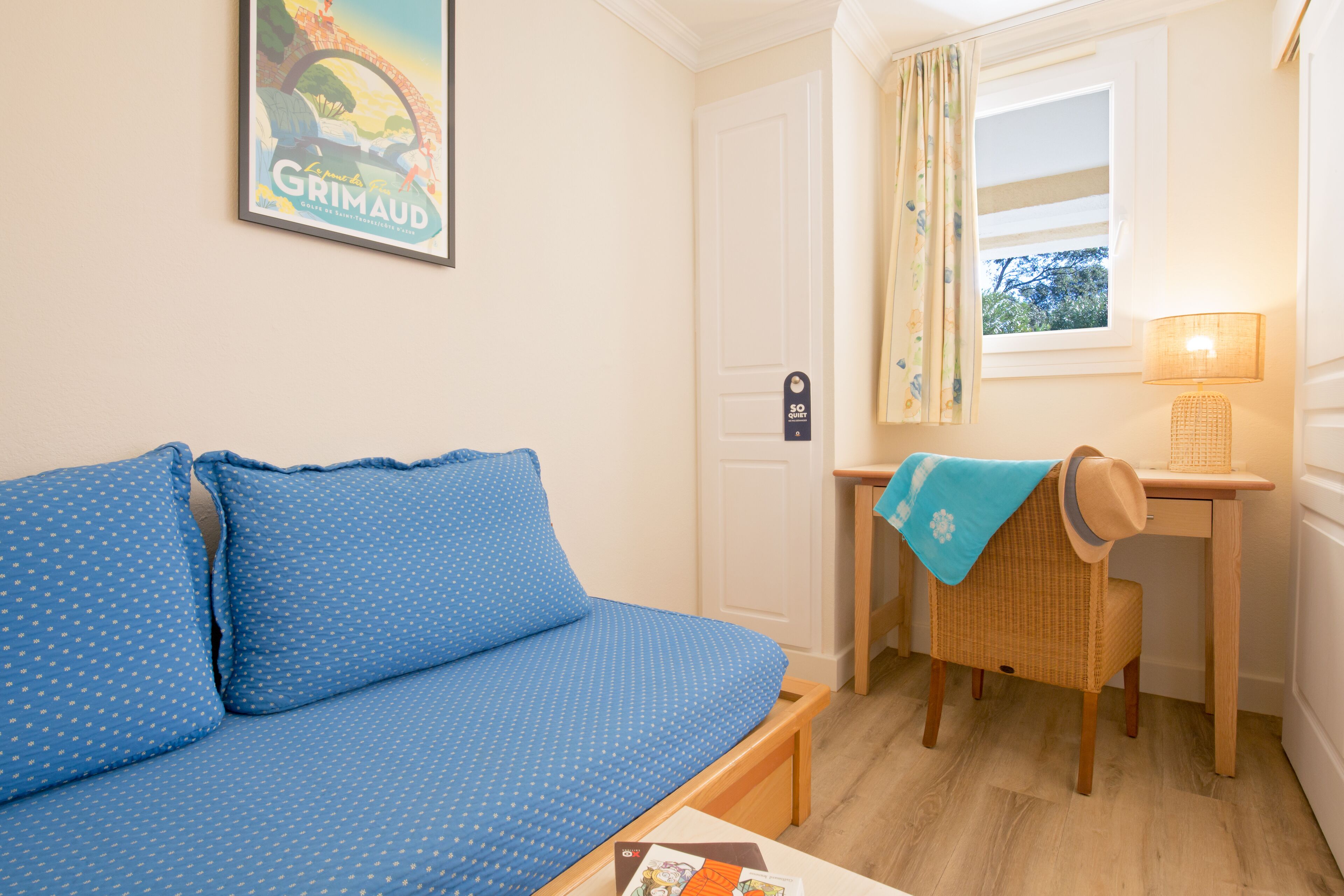 A room with a blue sofa, a small table with a lamp, a wicker chair, and a window with curtains.