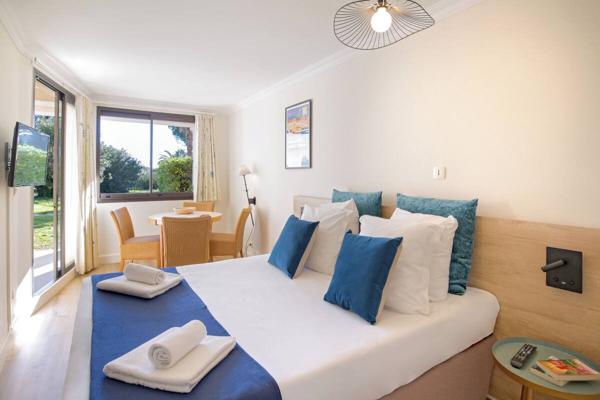 Ein Hotelzimmer mit einem großen Bett, einem Schreibtisch und einem Sessel. Ein Fenster bietet Blick auf grüne Natur.