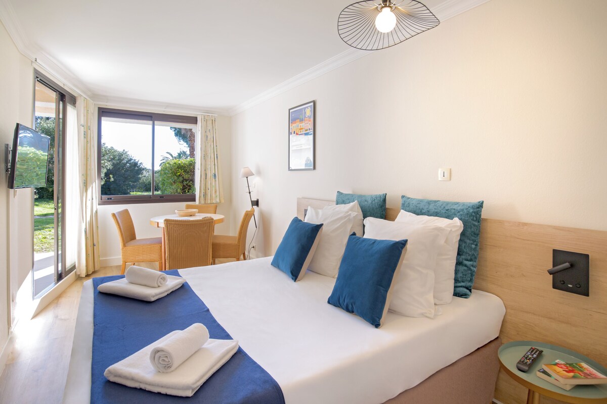 A hotel room with a large bed, a desk, and a chair. There is a window with a view of greenery outside.