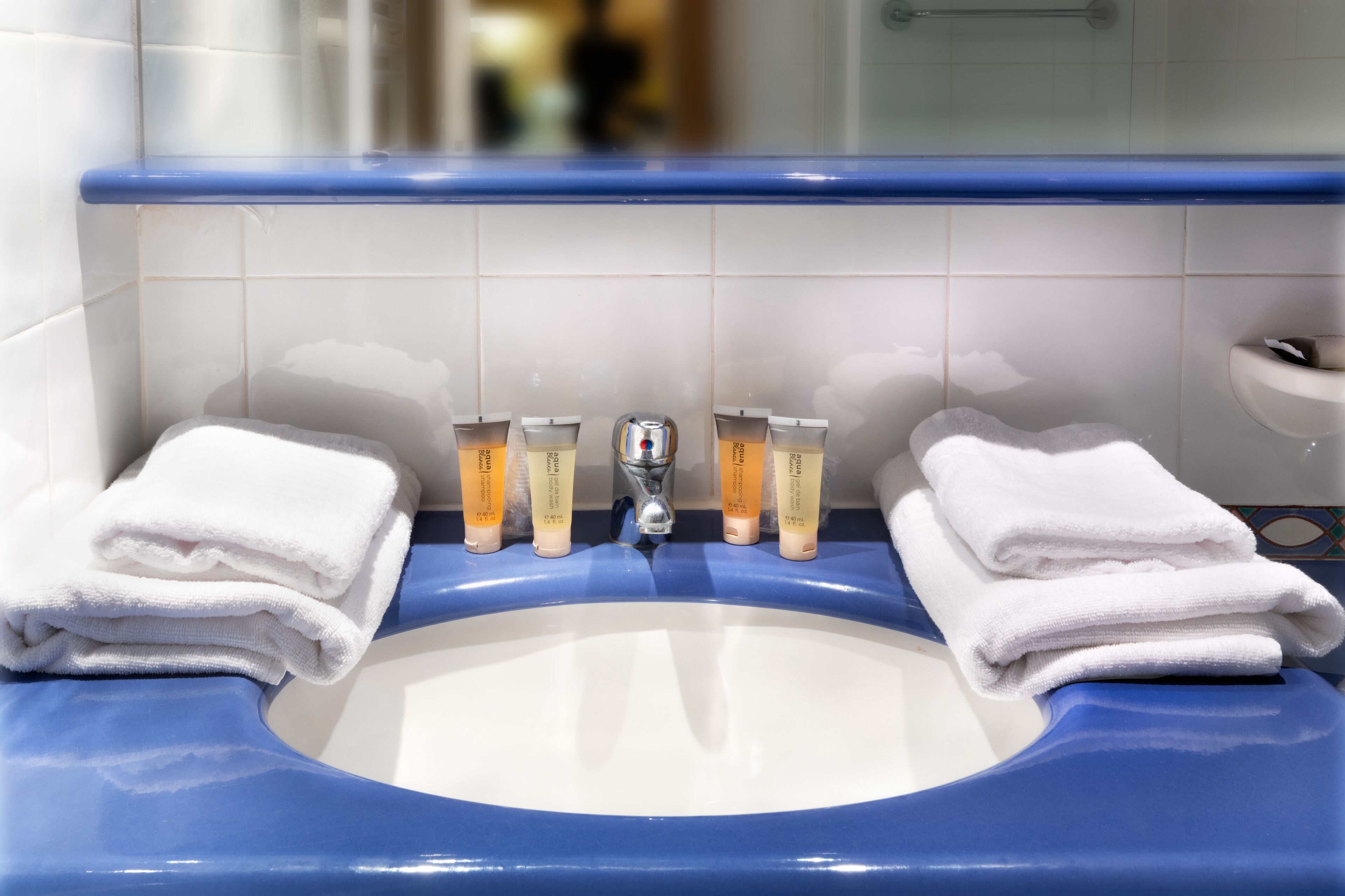 A bathroom sink with blue and white tiles, two white towels, and three orange tubes of lotion.