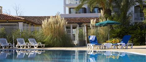 A swimming pool with lounge chairs and umbrellas, surrounded by palm trees and a building with balconies.