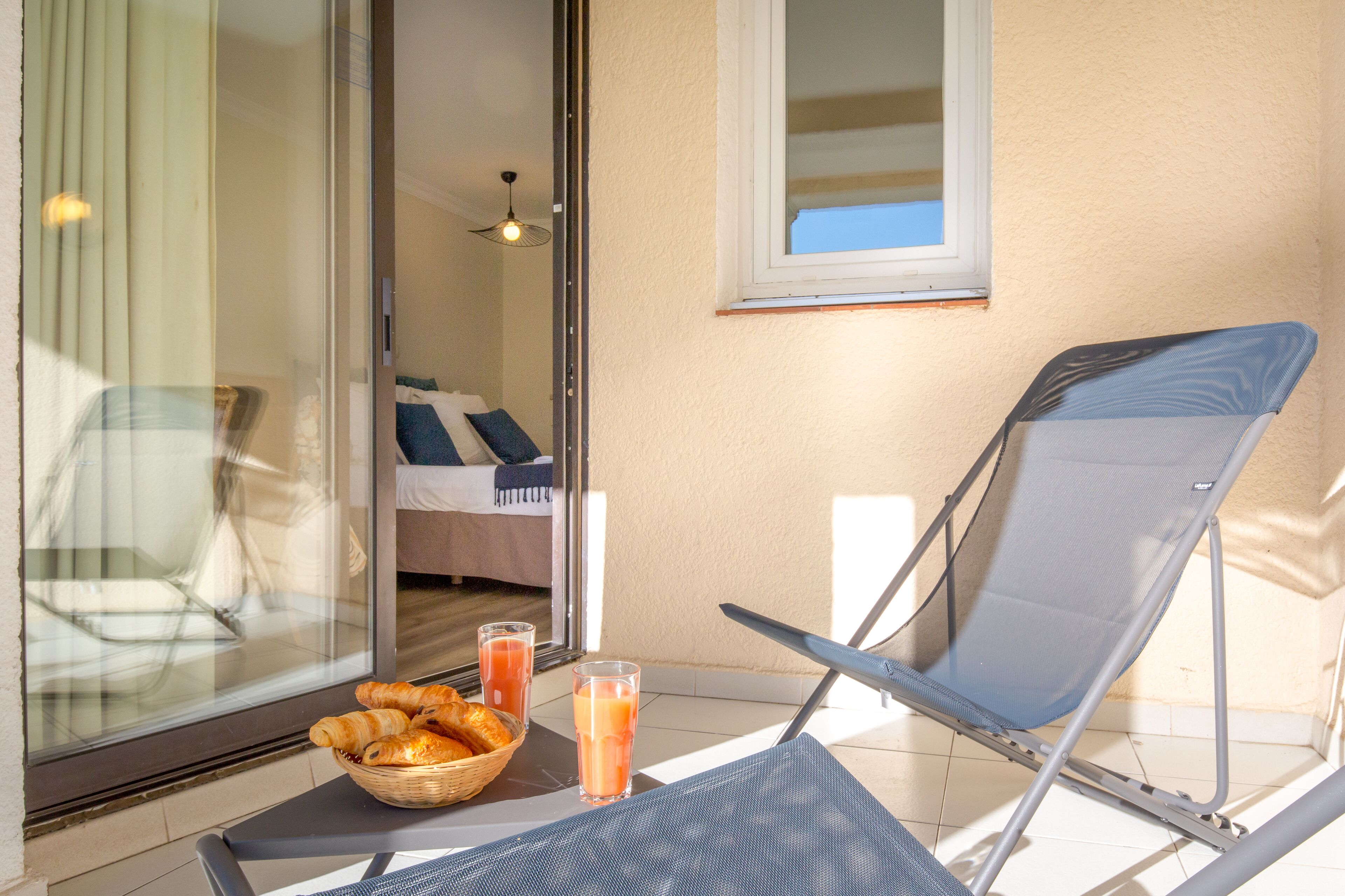 Un balcon avec une chaise, une table avec des croissants et du jus de fruits, et une vue sur l’intérieur.