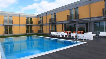 Un área de piscina moderna al aire libre, con una pileta rectangular, mobiliario blanco para exteriores y un edificio con balcones y ventanas amplias.