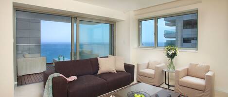 Una sala moderna con un sofá marrón, sillones beige, una mesa de centro con controles remotos y vistas al mar a través de amplias ventanas.