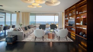 A modern living room with a large sofa, two armchairs, a coffee table, and a bookshelf. There is a large window with a view of the ocean. - Olympic Residence (Limassol)