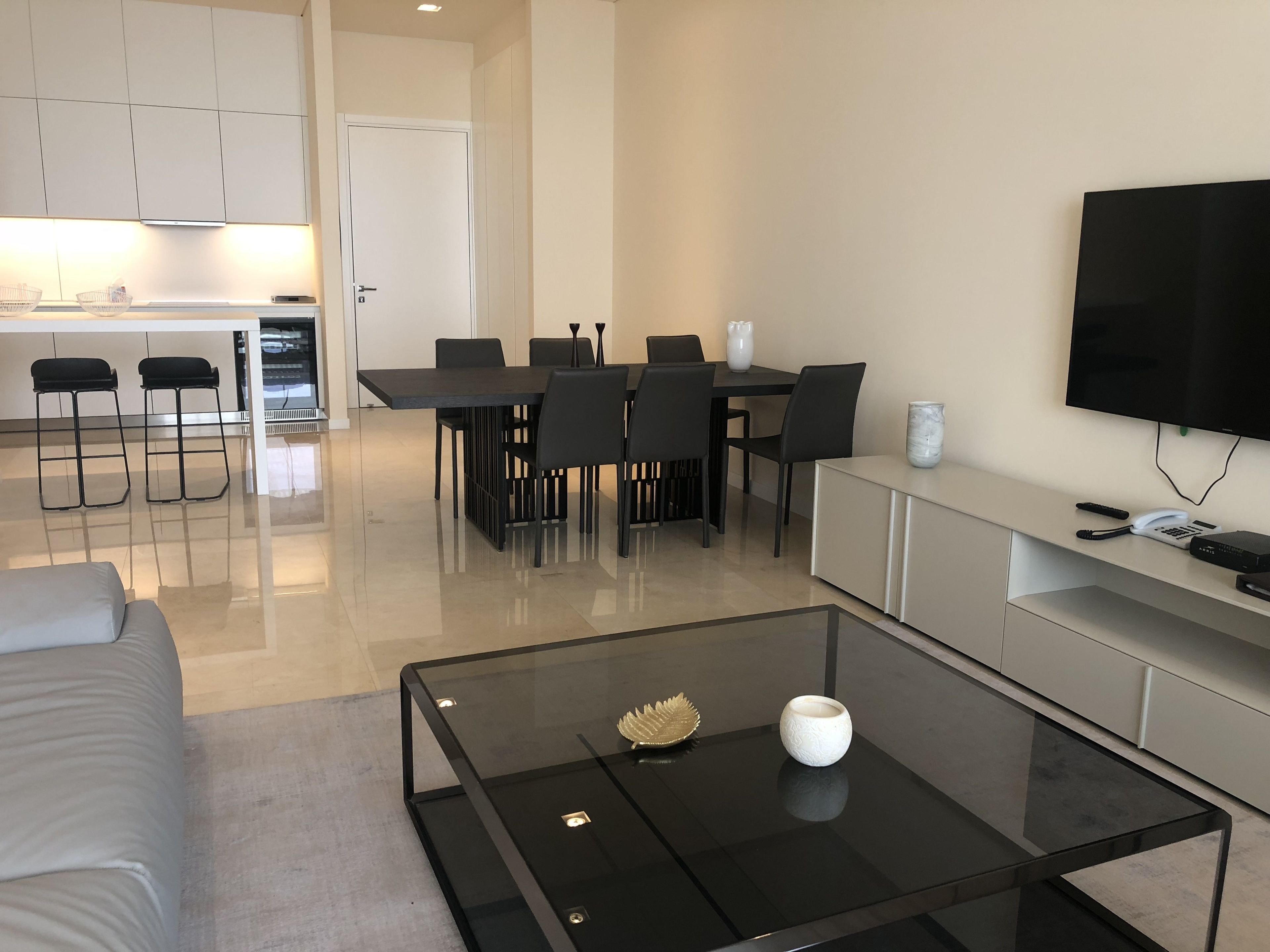 A modern living room with a glass coffee table, a sofa, a dining area with chairs, and a kitchen in the background.