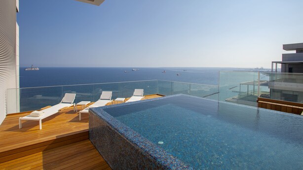 A modern infinity pool with lounge chairs, a glass railing, and a view of the ocean and ships. - Olympic Residence (Limassol)