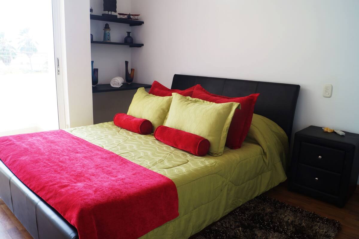 A bed with a red and yellow bedspread, two red pillows, and a black headboard. There is a small black nightstand with a drawer and a lamp on it.
