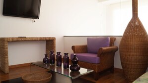 A living room with a flat-screen TV mounted on the wall, a wicker console table, a glass coffee table with purple vases, a purple armchair, and a large wicker vase. - Casa Blanca del Mar Hotel Boutique (Cartagena)