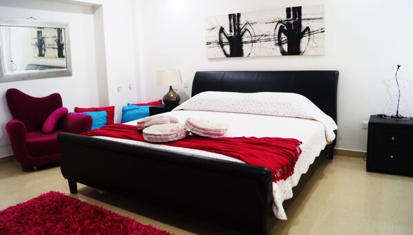 Un dormitorio con una cama negra, almohadas rojas y azules, una alfombra roja, una silla, una mesita de noche y una obra de arte en la pared.