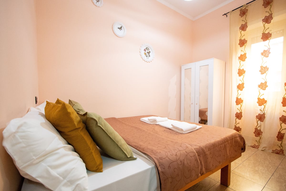 A neatly made bed with a brown bedspread and multiple pillows, a white door, and a floral curtain.