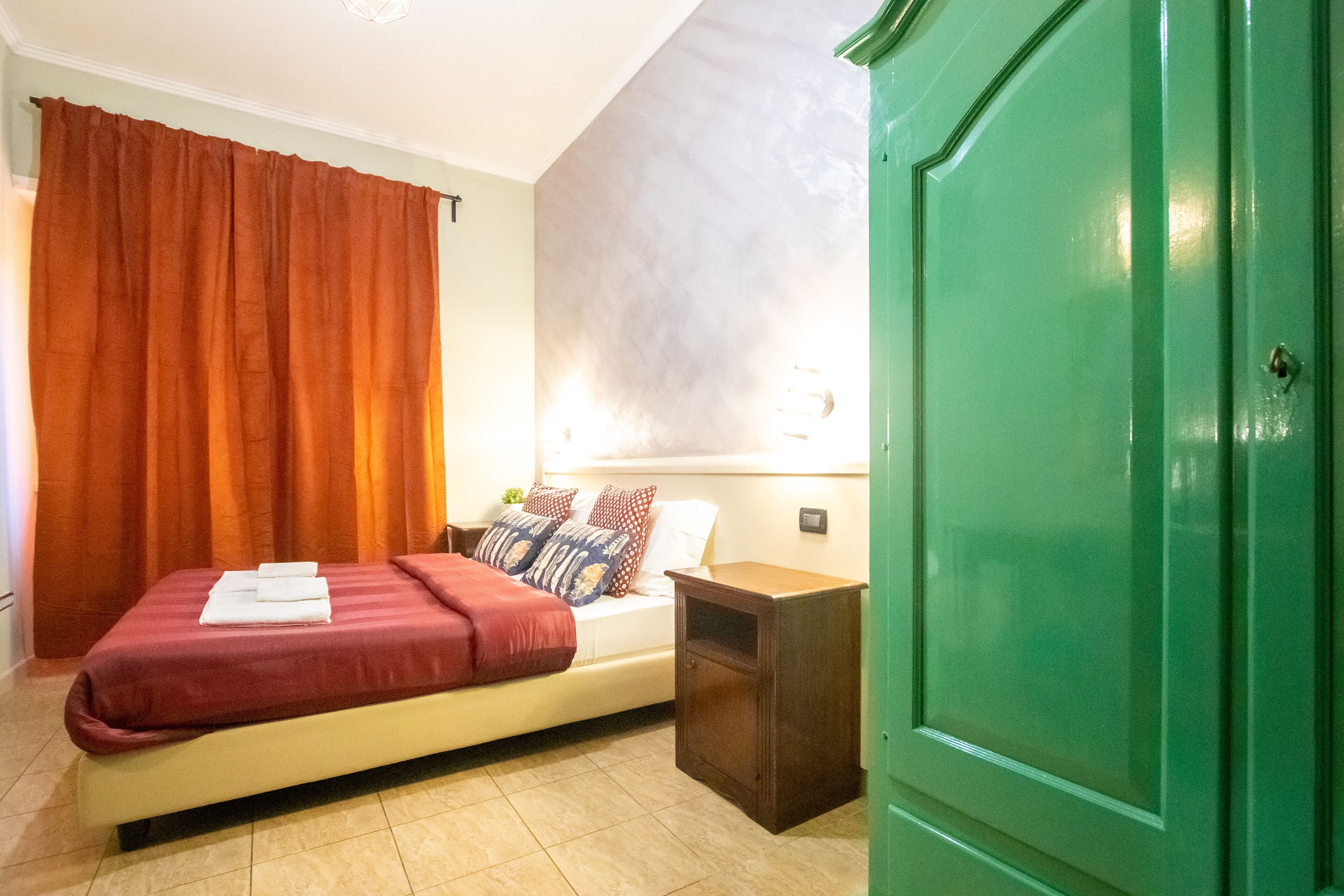 A bedroom with a bed, a nightstand, a green wardrobe, and a window with curtains.
