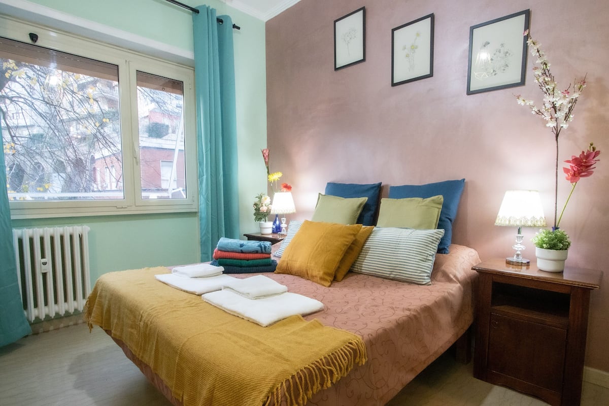 A bedroom with a bed, pillows, a nightstand, a window, and a radiator.