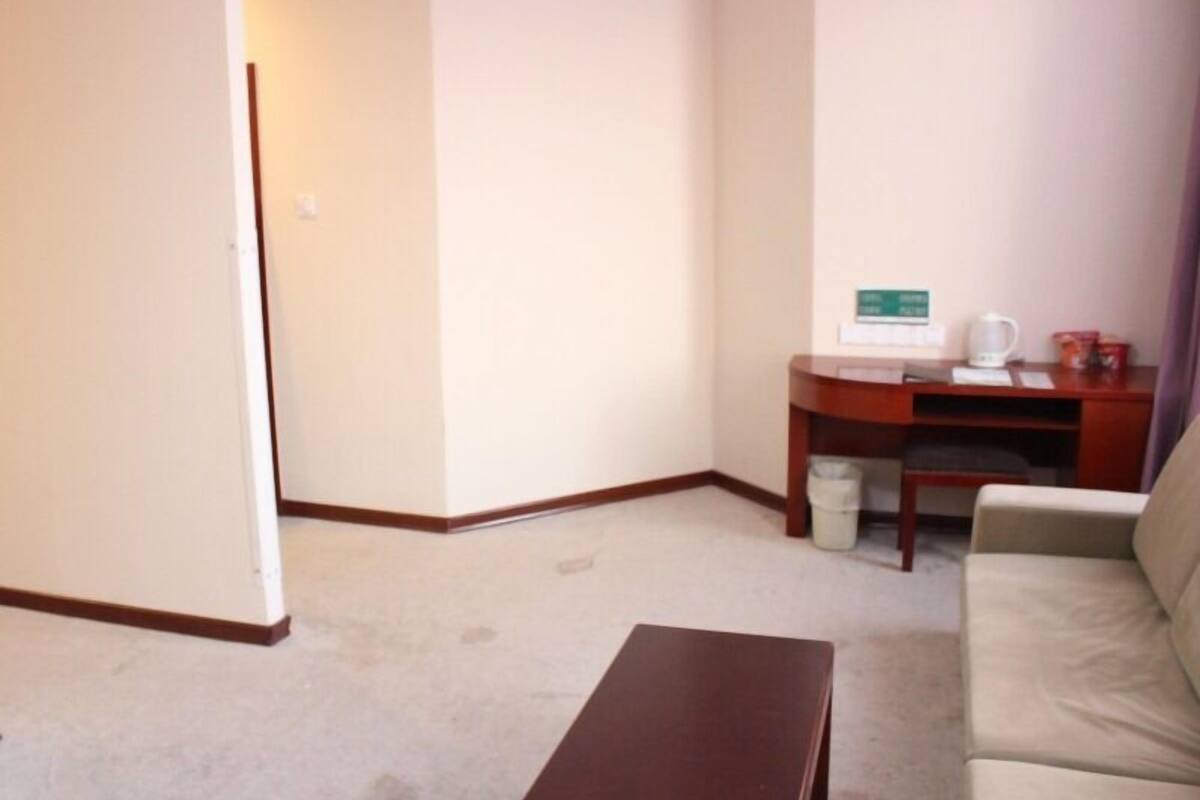 A hotel room with a beige sofa, a wooden coffee table, a desk with a computer, and a trash bin.