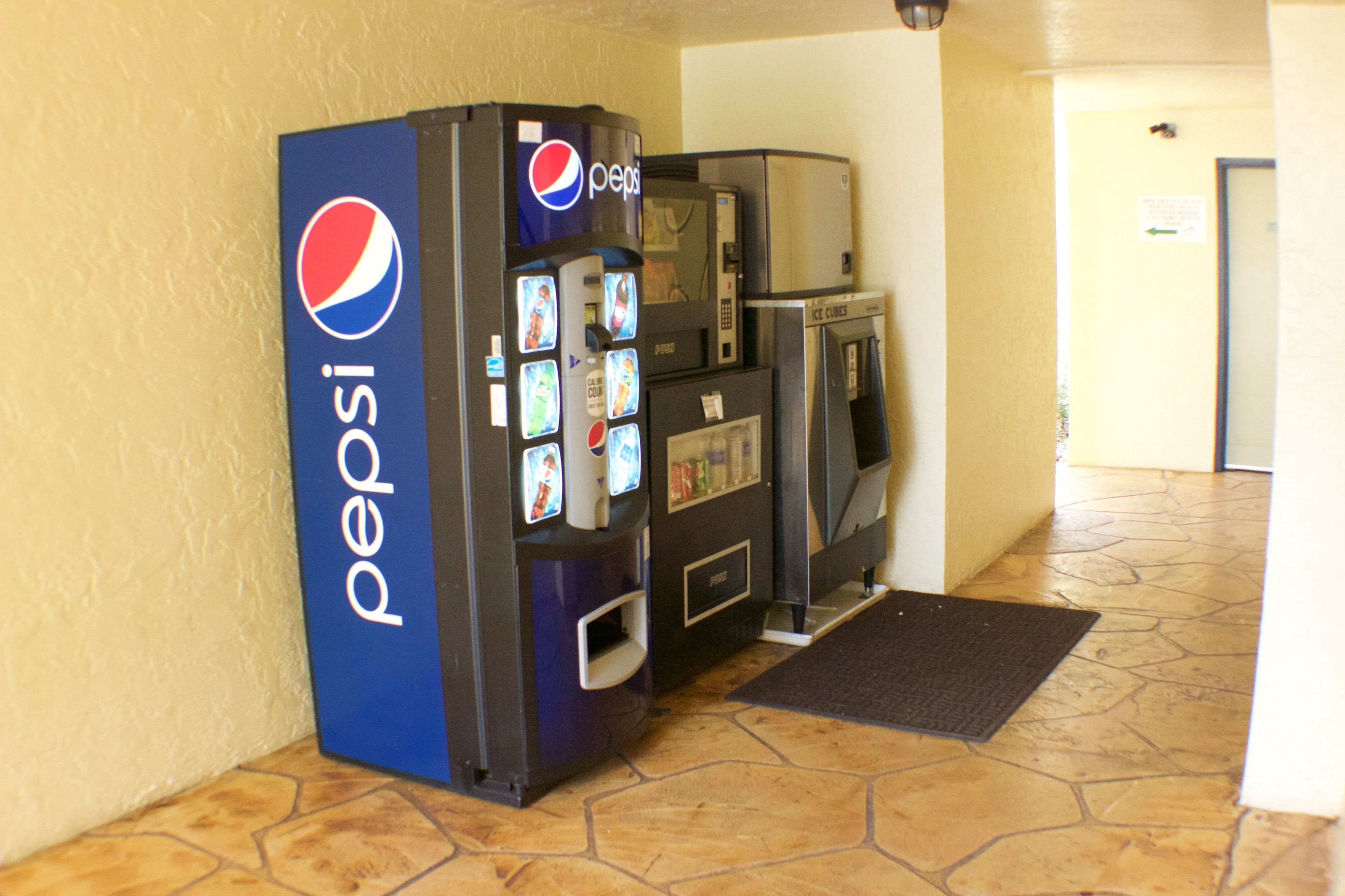 A Pepsi vending machine and a microwave in a hallway with tiled flooring.