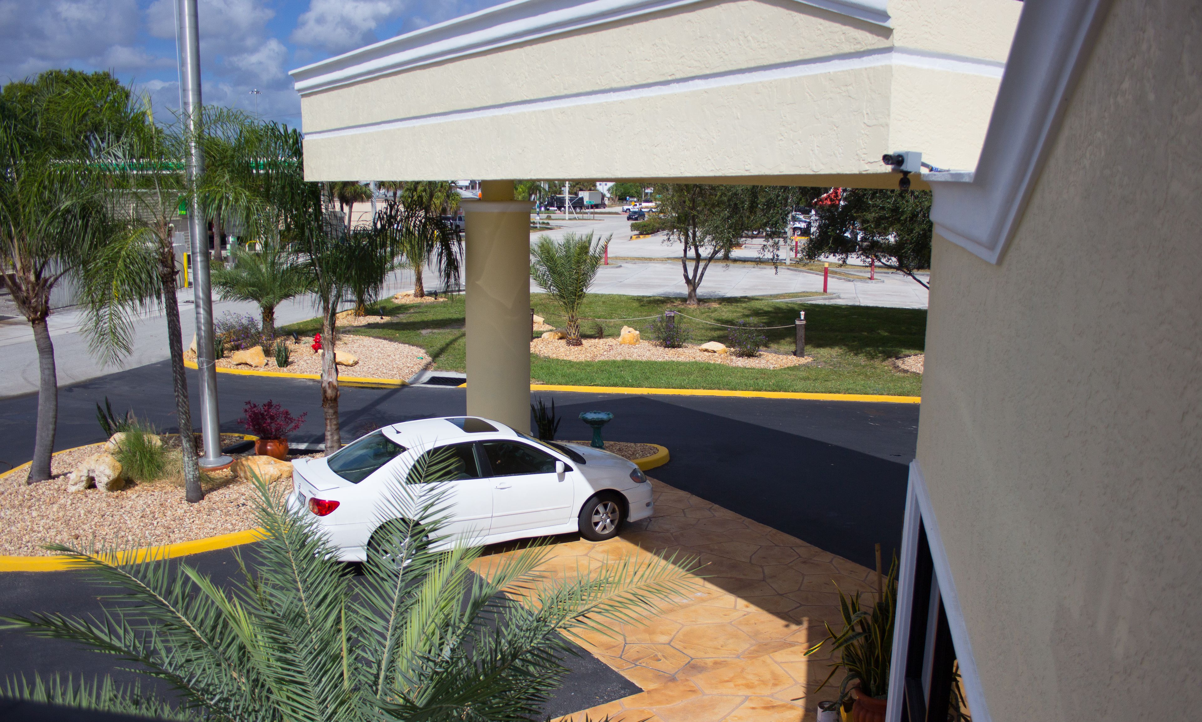 A white car parked under a covered area with a stone patio, palm trees, and a view of a street with a traffic light and vehicles.