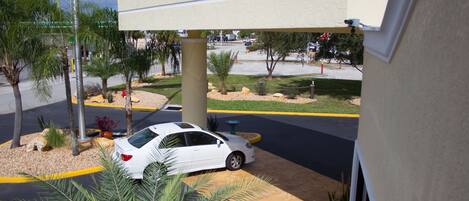 A white car parked under a covered area with a stone patio, palm trees, and a view of a street with a traffic light and vehicles.