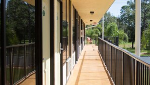 A covered corridor with wooden flooring, black railings, and glass doors leading to rooms.