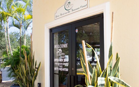 A hotel entrance with a welcome sign, a black mat, and two potted plants.