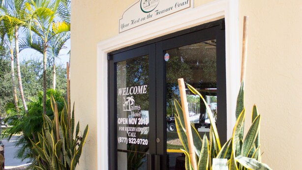 A hotel entrance with a welcome sign, a black mat, and two potted plants.