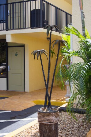 A building with a yellow exterior, a black metal balcony railing, and a palm tree sculpture in the foreground.