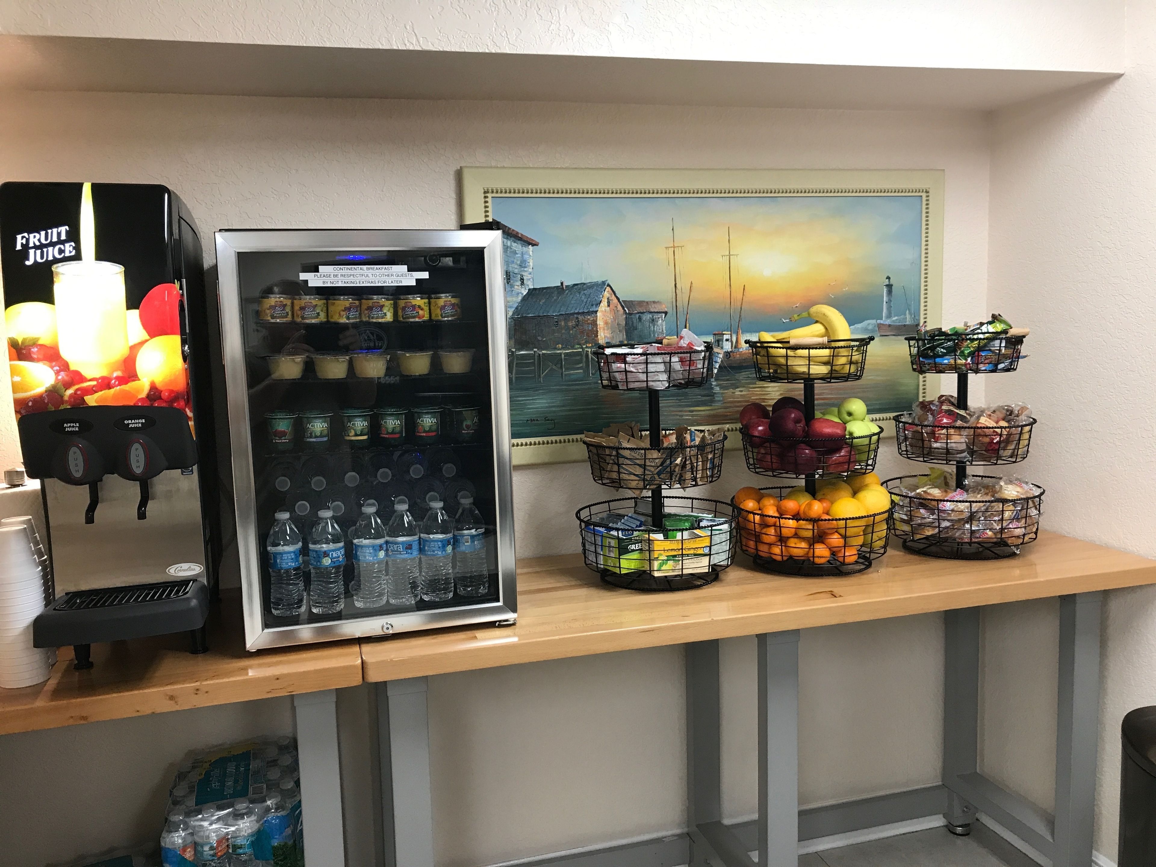 A refreshment station with a fruit juice dispenser, a refrigerator stocked with beverages, and baskets of fruits and snacks.