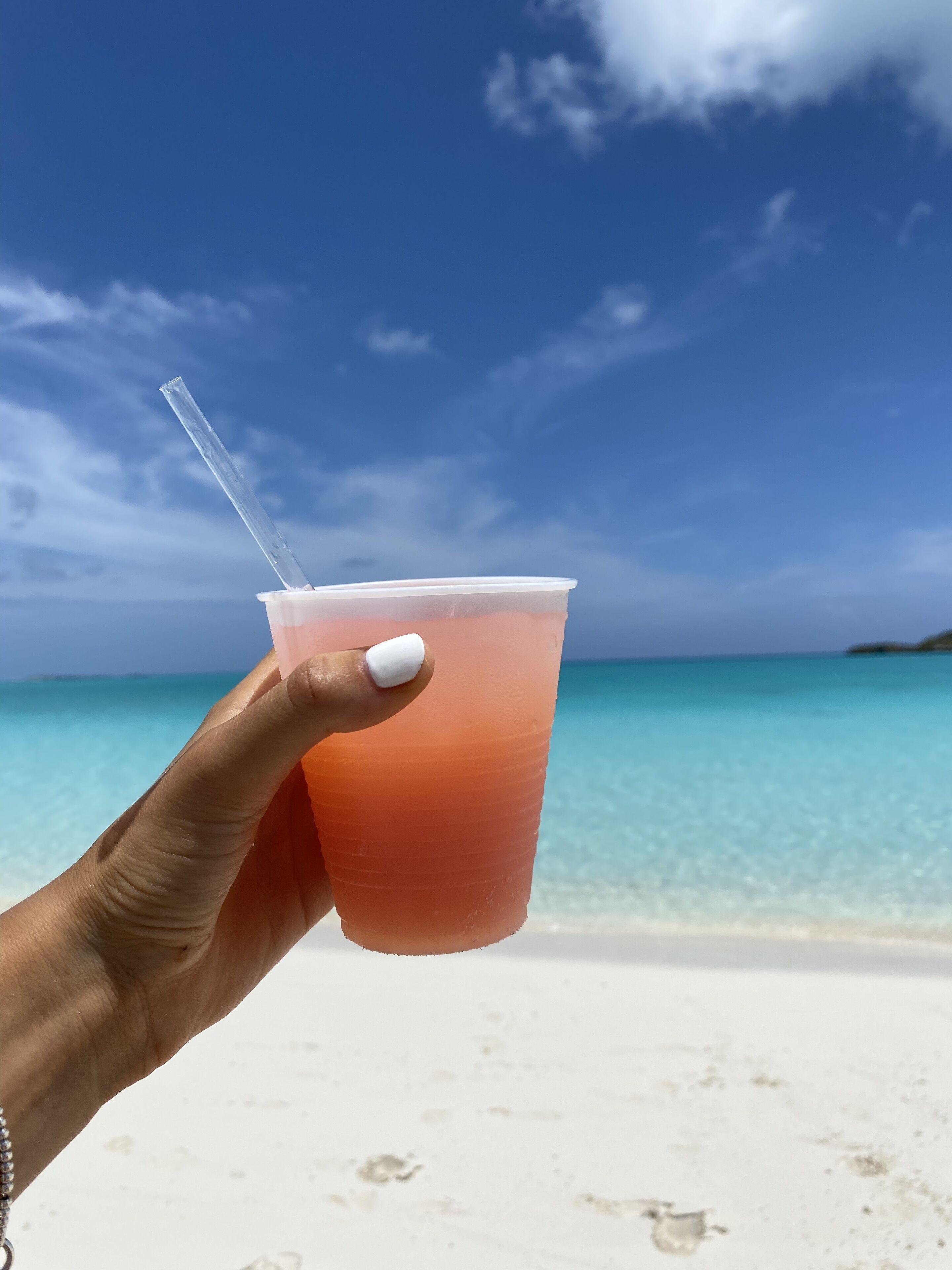 Een hand die een plastic beker met rietje vasthoudt, handpalm is wit geschilderd, met op de achtergrond een strand en helder turquoise water en een heldere lucht.
