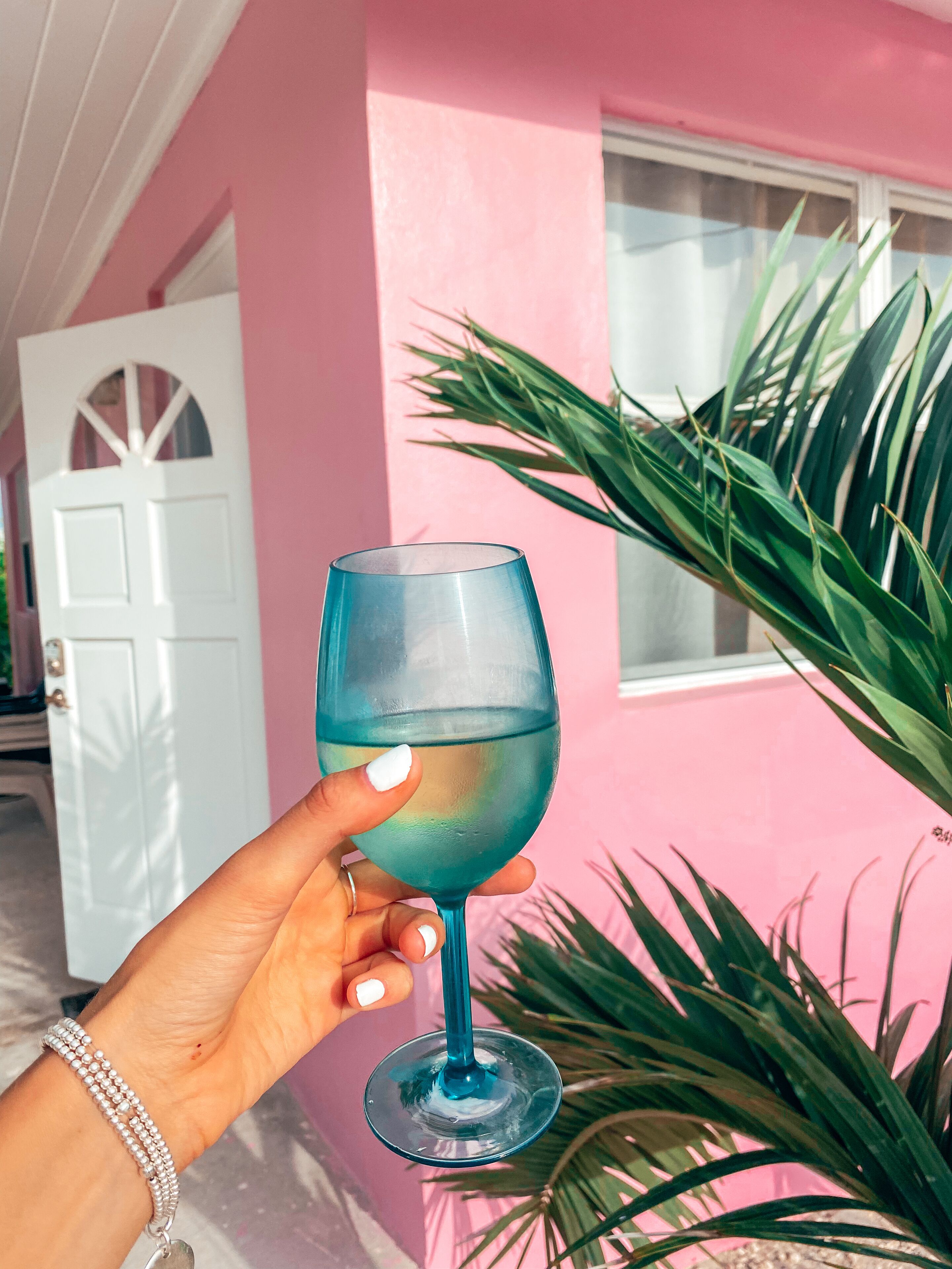 A hand holding a clear glass with a blue tint, palm tree, and a pink building with a white door in the background.