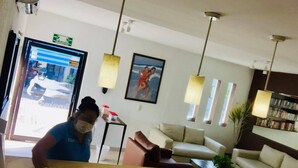 A hotel reception area with a receptionist, a reception desk, a seating area with couches, pendant lights, a framed picture on the wall, and a bookshelf. - Suites Princess (Bucerías)