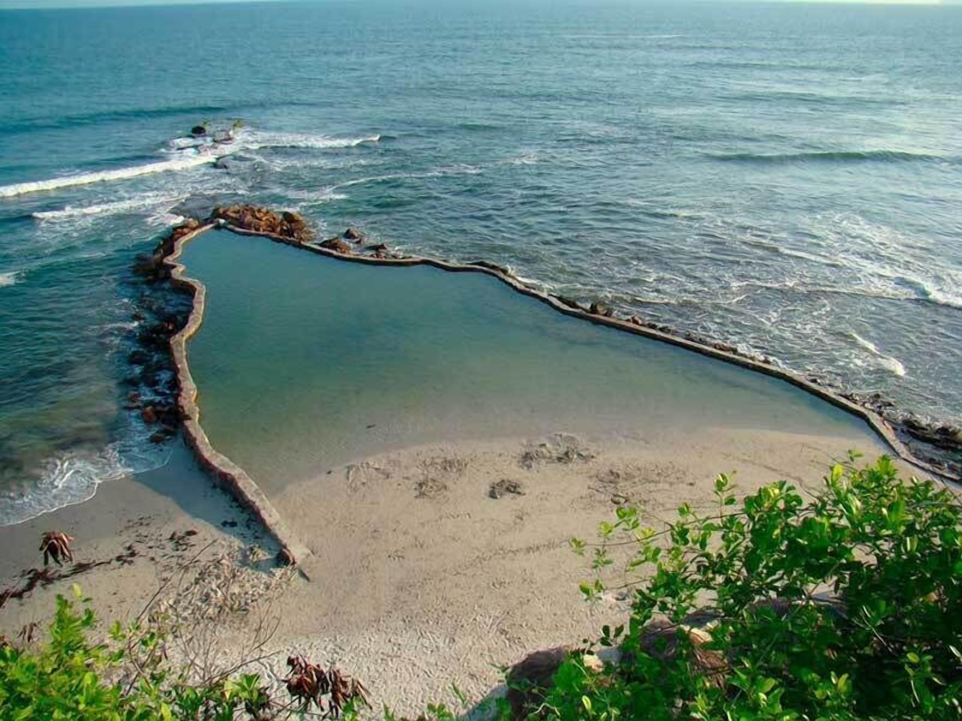 Ein Küstenstrand mit klarem Wasser, felsigem Ufer und spärlicher Vegetation.