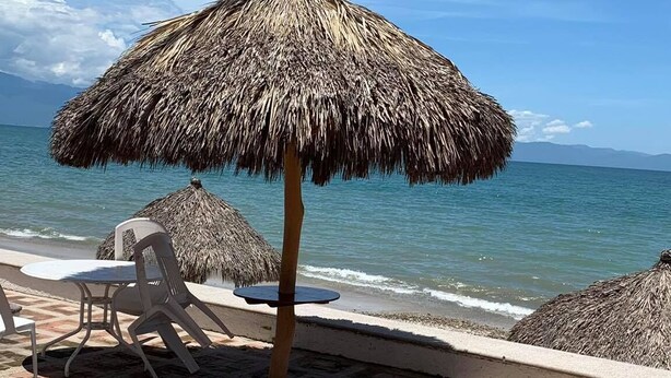 A beachside patio with a thatched umbrella, white plastic chairs, and a view of the ocean. - Suites Princess (Bucerías)