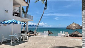 A beachfront patio with white plastic chairs and tables, striped blue and white umbrellas, and a thatched umbrella. - Suites Princess (Bucerías)
