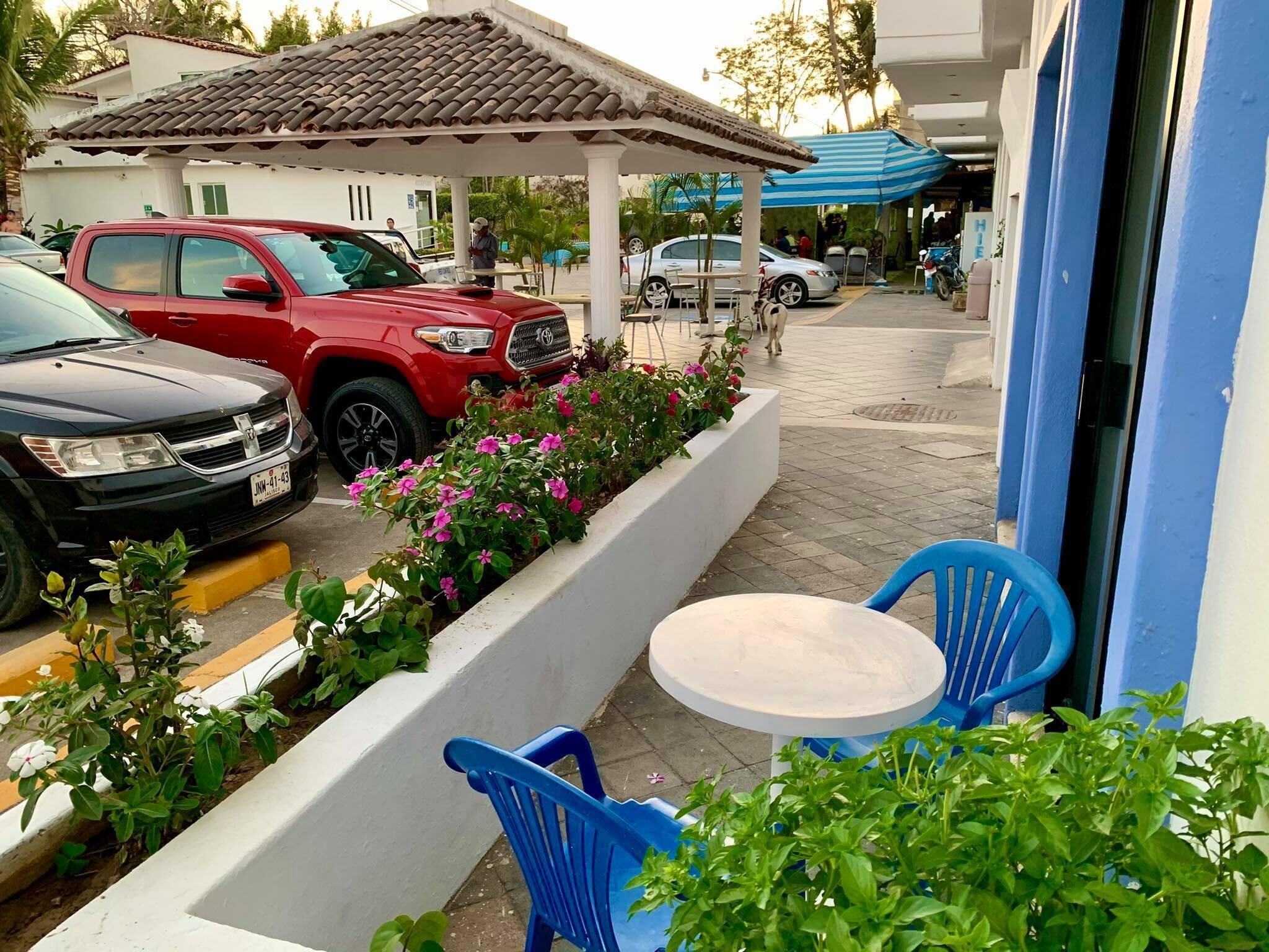 A parking area with a red SUV and a black car, a small outdoor seating area with blue chairs, and a white building with a tiled roof.