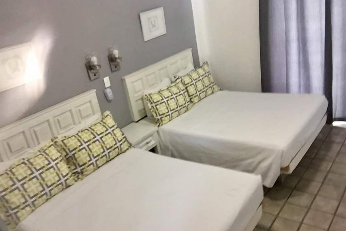 Habitación de hotel con dos camas individuales, cada una con colchón blanco y almohadas estampadas. Entre las camas hay una mesita de noche con una lámpara y un teléfono. La pared es gris y hay una ventana con cortinas.