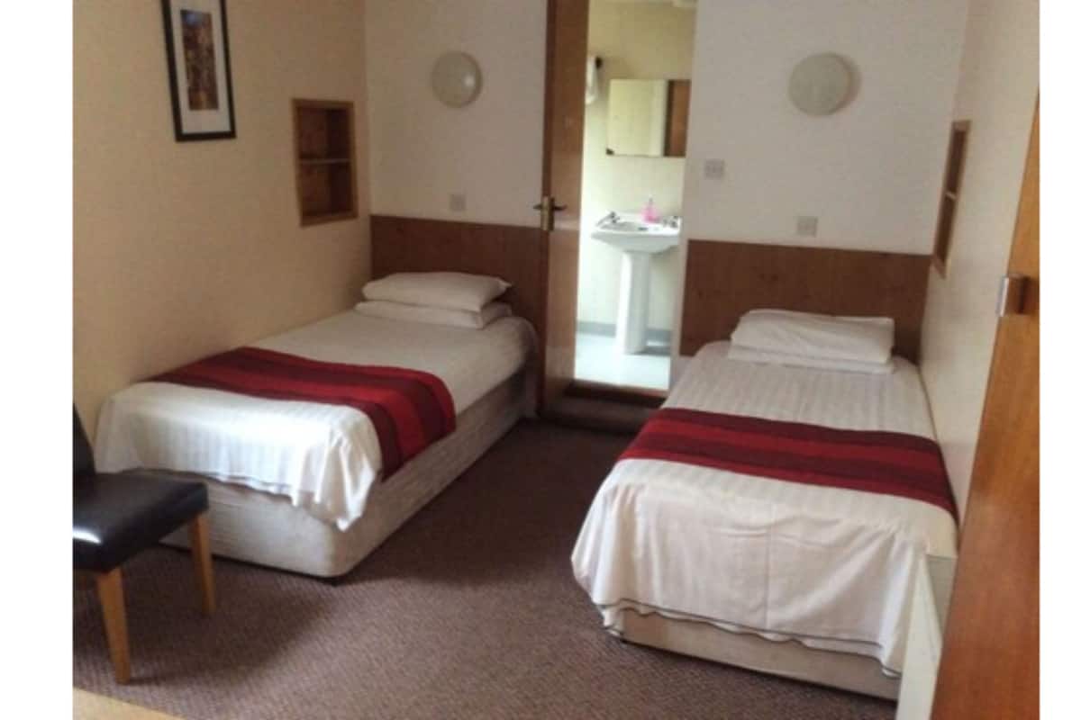 A small, clean room with two single beds, a chair, a small shelf, and a bathroom visible through a door.