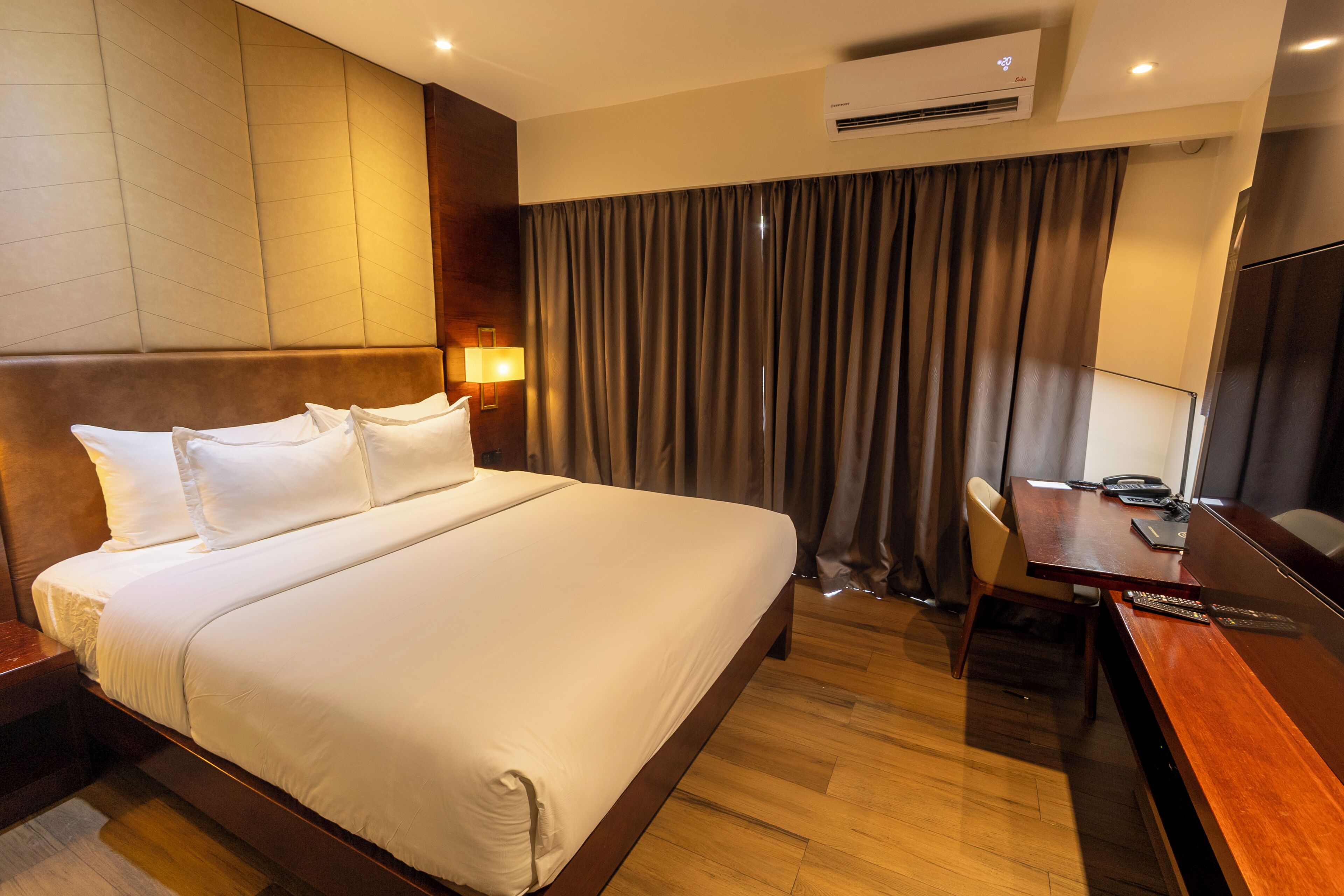 A hotel room with a large bed, a desk, a chair, an air conditioning unit, and a curtain.