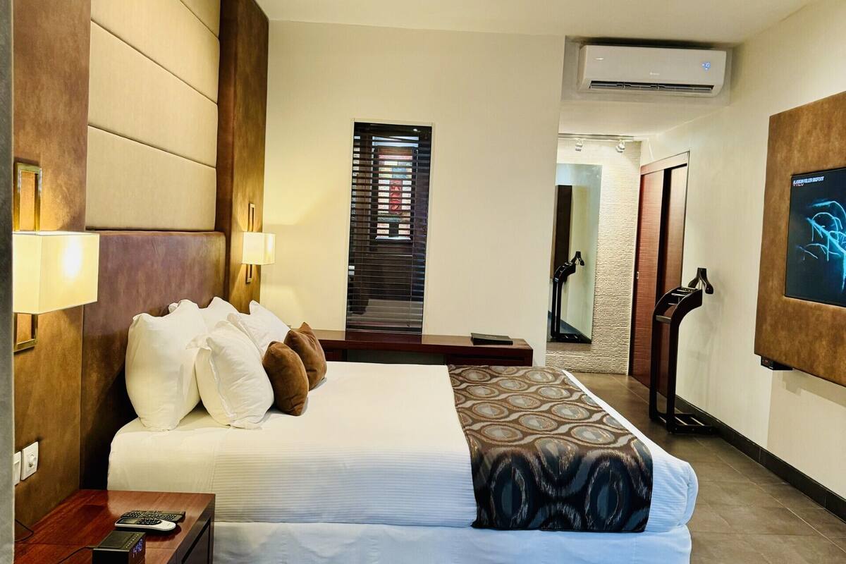 A hotel room with a large bed, bedside tables, a TV, an air conditioning unit, and a door leading to another room.