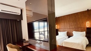 Habitación de hotel moderna con televisión de pantalla plana montada en una consola de madera, un escritorio con una silla, una cama con ropa de cama blanca y un ventanal con vistas.