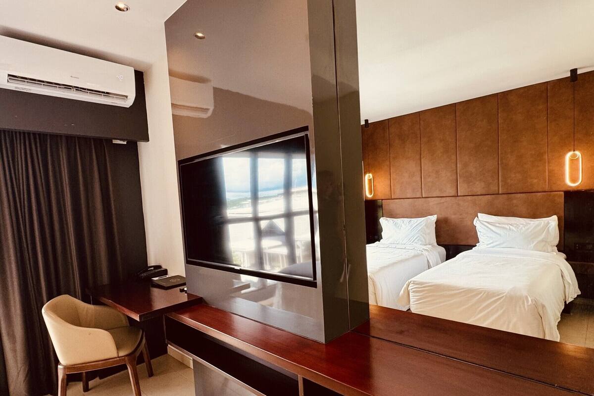A modern hotel room with a flat-screen TV mounted on a wooden console, a desk with a chair, a bed with white linens, and a large window offering a view.