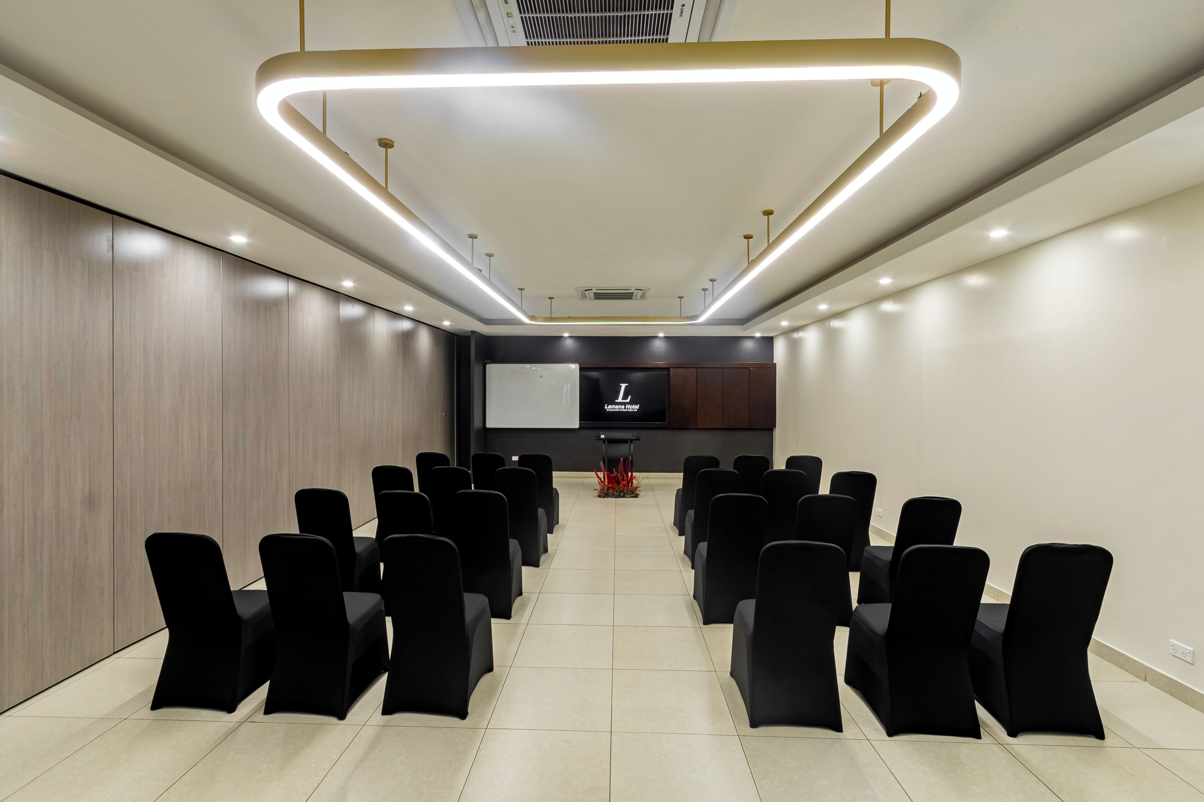 A conference room with rows of black chairs, a projector screen, and a modern ceiling light fixture.