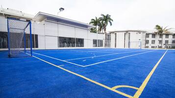 An outdoor basketball court with a blue surface and yellow lines, surrounded by buildings and palm trees.