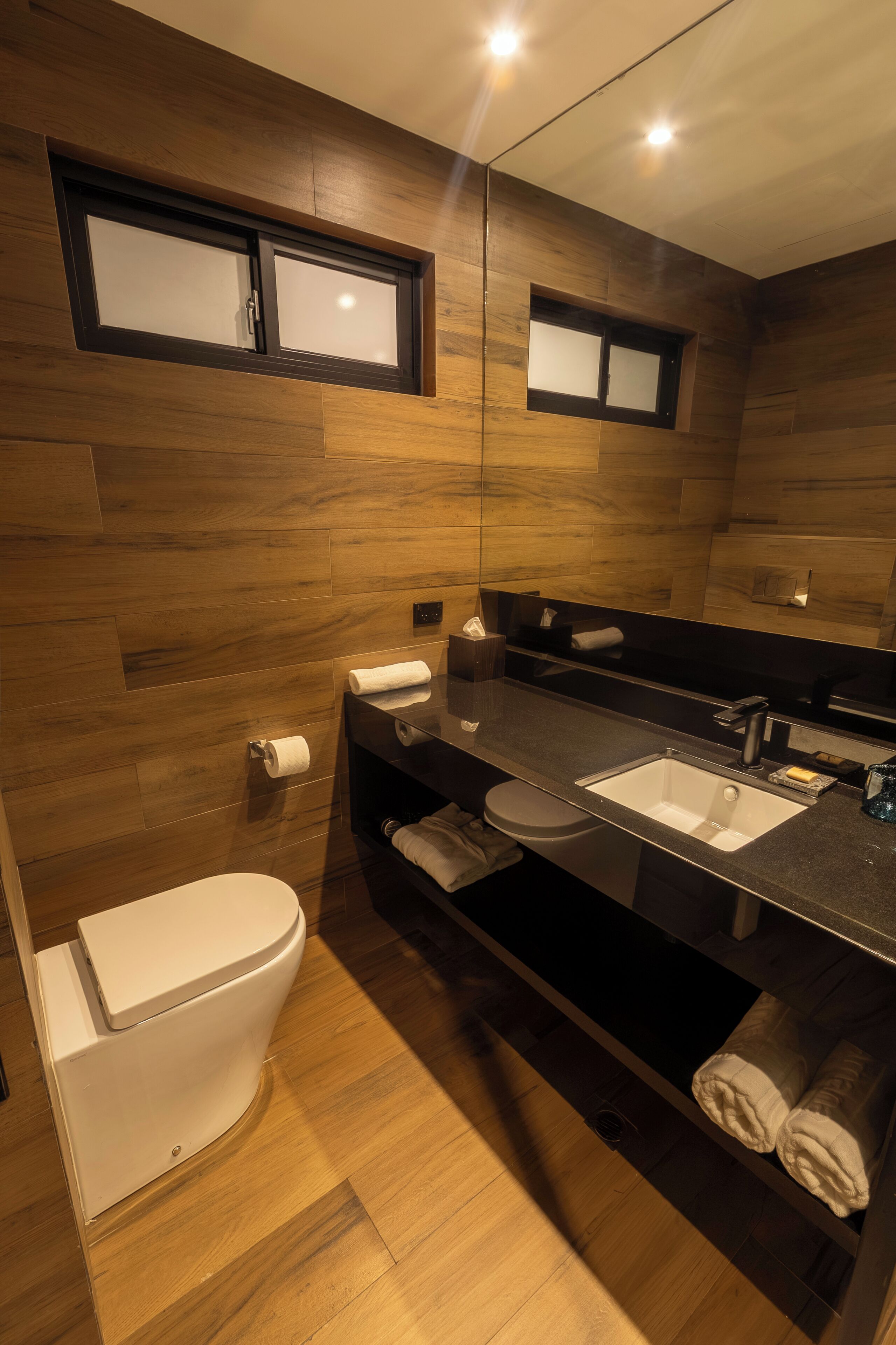 Une salle de bain moderne avec un plancher en bois, des toilettes blanches, un lavabo noir et un miroir au-dessus du lavabo.