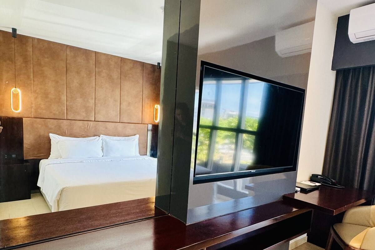 A modern hotel room with a large flat-screen TV, a wooden desk, a comfortable bed, and a view of greenery outside.