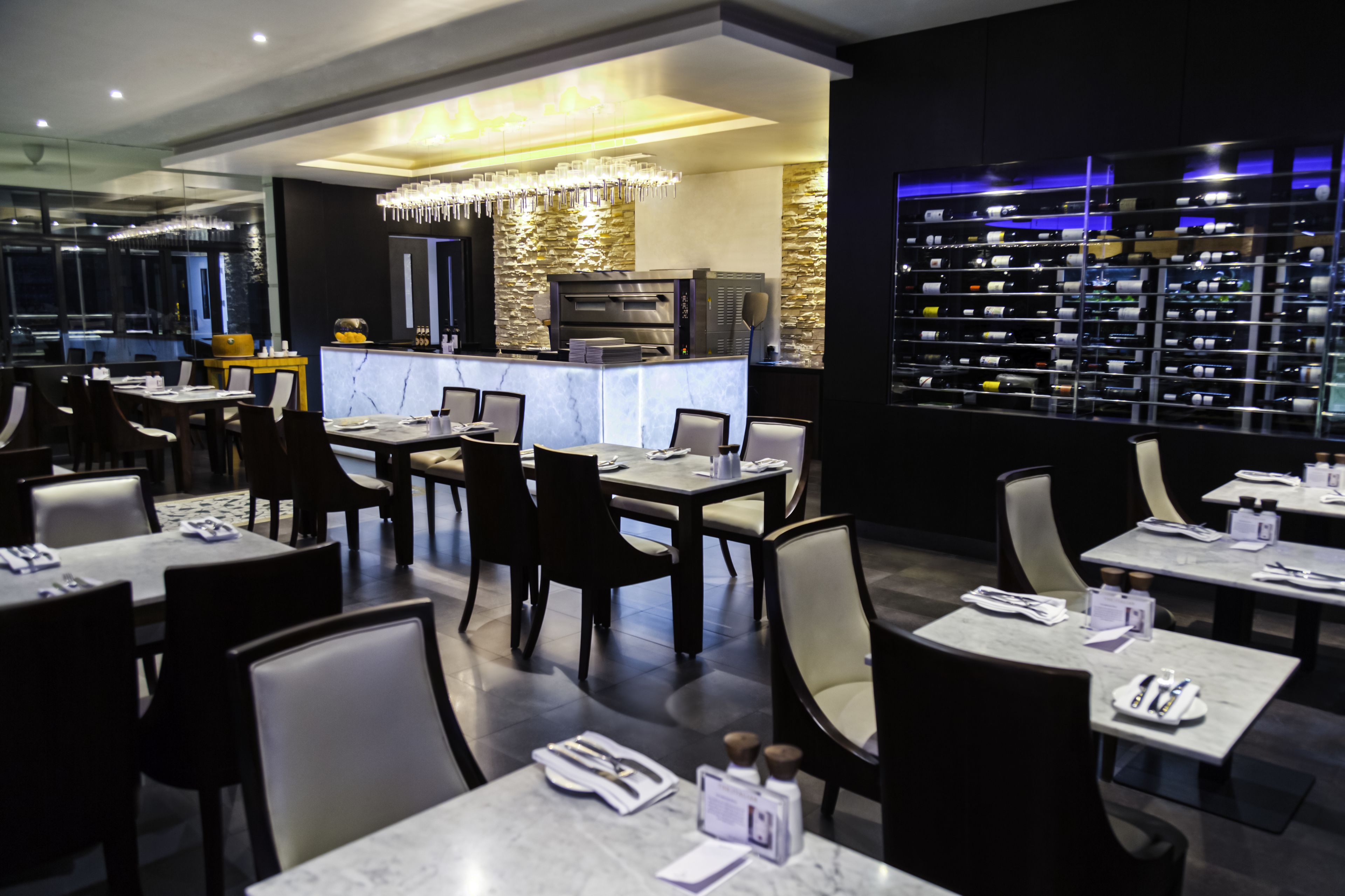 A modern restaurant with marble tables, black and white chairs, a bar area with a wine rack, and a large wine bottle display.
