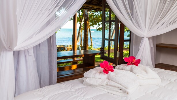 A bed with white linens and a pink flower, a wooden bench, and a view of the sea through a window with white curtains. - Enseada do Espelho (Porto Seguro)