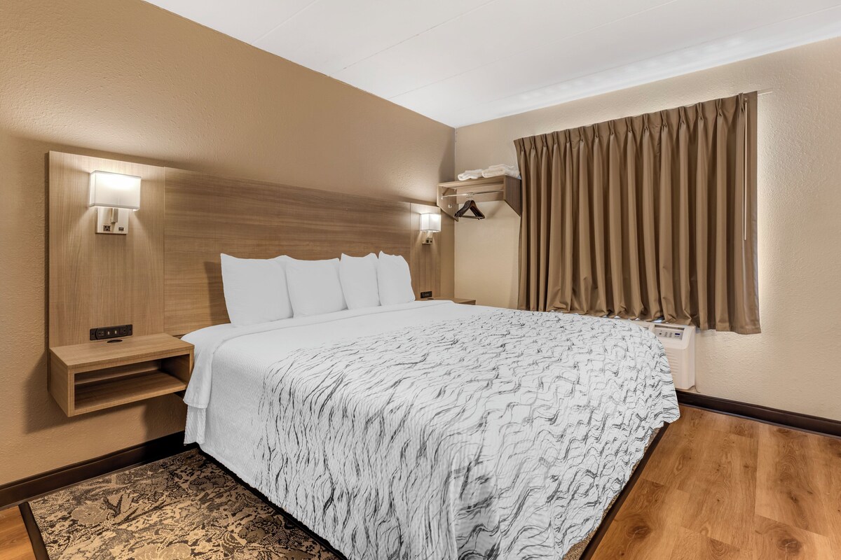 A hotel room with a large bed, two bedside tables, a wall-mounted light, a window with curtains, and a wooden floor.