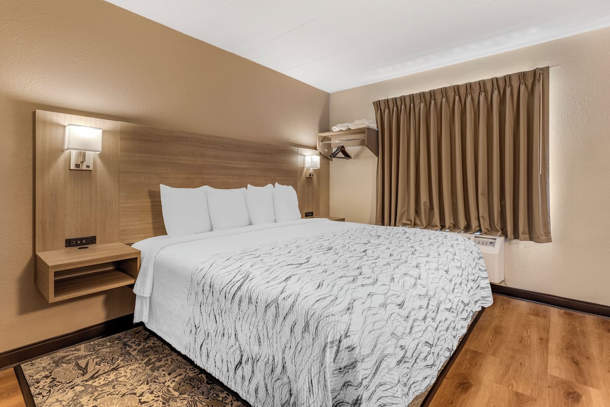 A hotel room with a large bed, two bedside tables, a wall-mounted light, a window with curtains, and a wooden floor.