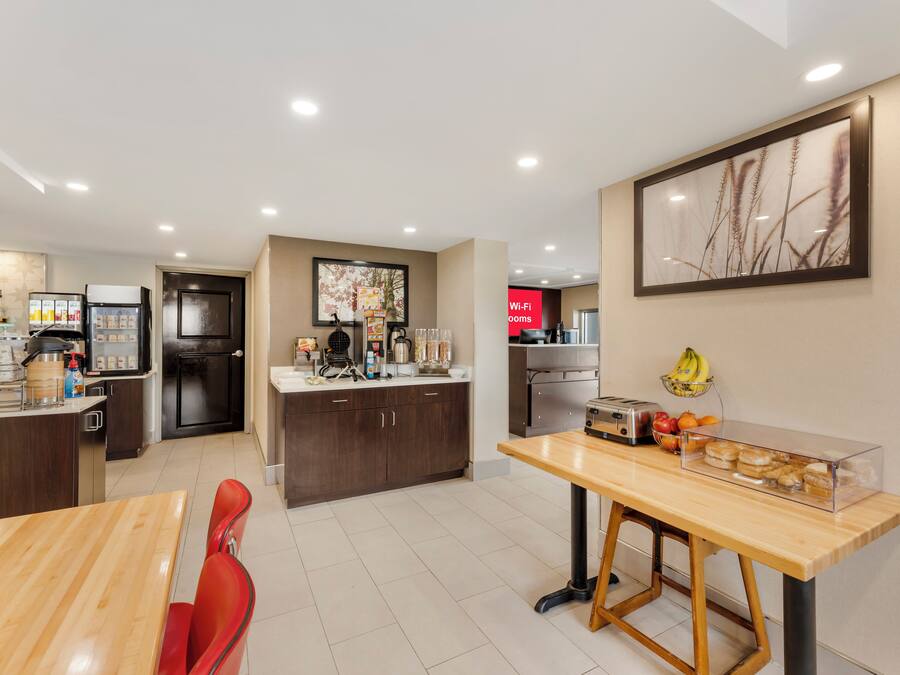 A modern kitchen with a central island, a dining area with a wooden table and red chairs, and a kitchen counter with various appliances and a fruit basket.