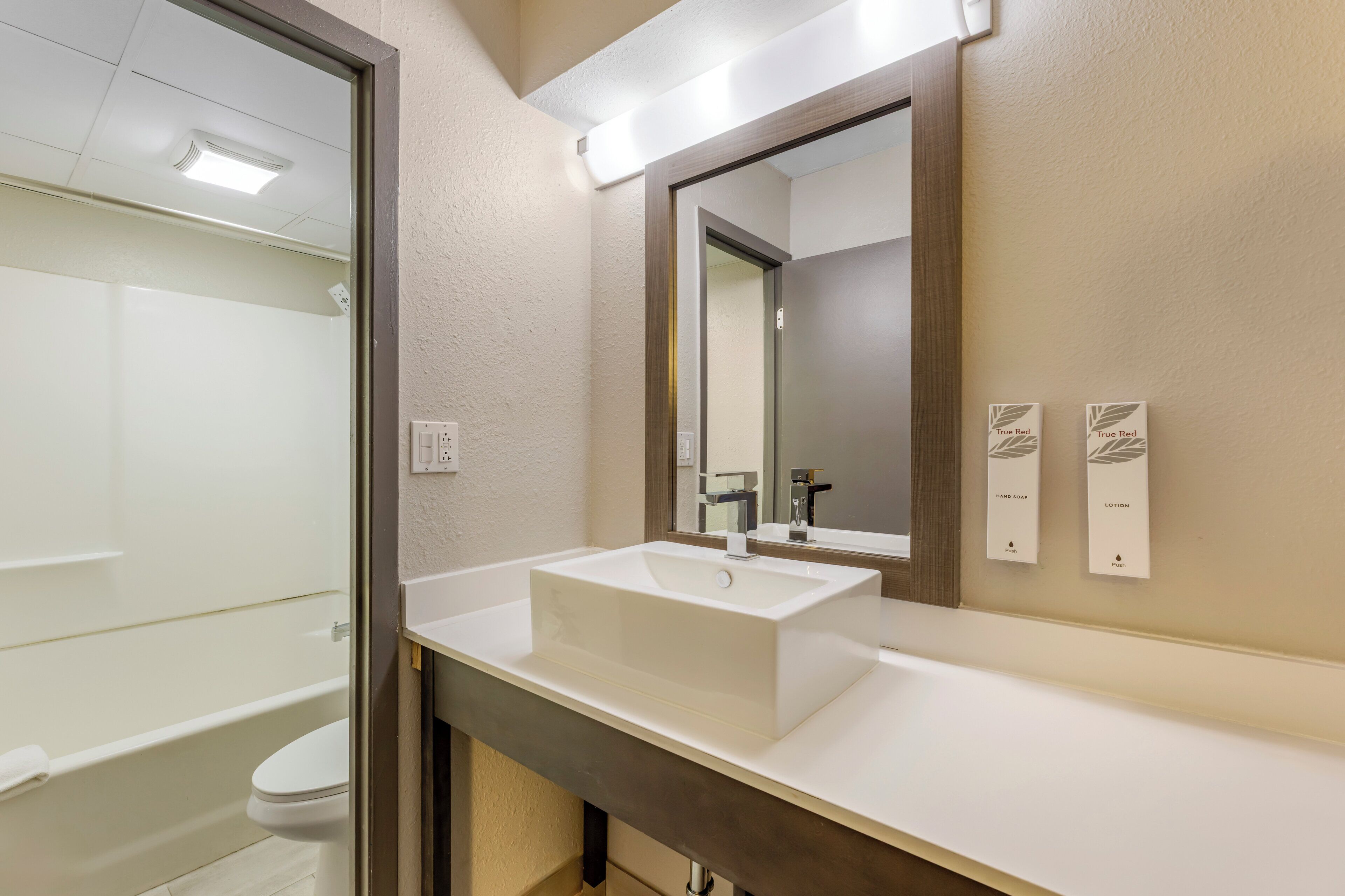 A bathroom with a white sink, a mirror, and a toilet. There are two wall-mounted soap dispensers labeled 'True Red Hand Soap' and 'True Red Lotion.'